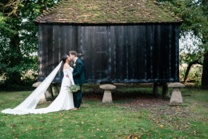 A bride and groom share a kiss outside, standing in front of a rustic wooden structure elevated on stone supports. The bride wears a long white gown with a veil, and holds a bouquet, while the groom is in a dark suit. Lush greenery surrounds them.