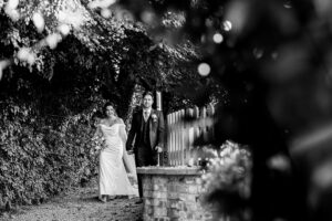 A black and white photo of a bride and groom walking on a path surrounded by trees and foliage. The bride is wearing a long dress, and the groom is in a suit. They appear joyful and are holding hands.