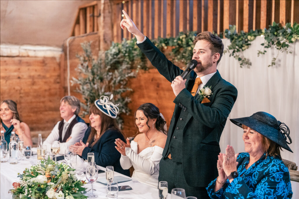 A man in a suit, holding a microphone, gestures while giving a speech at a wedding reception. He stands at a table with smiling guests, including women in hats and formal attire, set with floral arrangements and glasses.