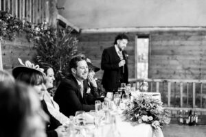 A black-and-white photo of a wedding reception. Guests, including a man and woman seated at a long table, listen to a man in a suit giving a speech with a microphone. The table is adorned with flowers and glasses.