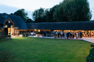 A large barn-like building with string lights hosts a gathering at dusk. People are socializing outside on a paved area surrounded by a lush green lawn. Trees are visible in the background under a slightly overcast sky.