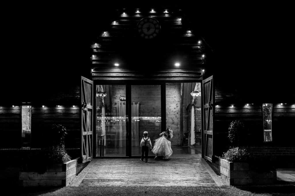 Two children run joyfully out of a large, illuminated barn at night. The barn doors are wide open, and the building is softly lit with glowing lights, creating a warm and inviting atmosphere.