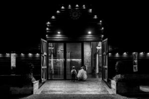 Two children run joyfully out of a large, illuminated barn at night. The barn doors are wide open, and the building is softly lit with glowing lights, creating a warm and inviting atmosphere.