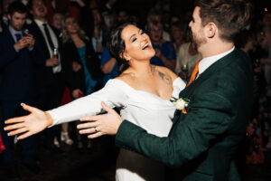 A bride and groom joyfully dance at their wedding reception. The bride is wearing a white dress and has her arms outstretched, while the groom in a dark suit smiles at her. Guests in the background watch and celebrate.