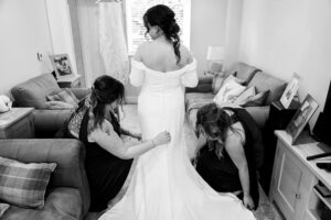 A bride, seen from the back, stands in a living room wearing an off-the-shoulder wedding gown. Two women assist her with the dress train. The room is furnished with a sofa, pillows, and framed photos, creating a cozy atmosphere.