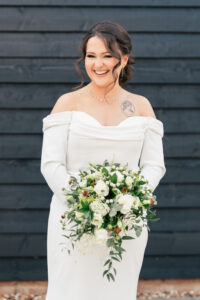 Bride in an off-the-shoulder white dress holding a bouquet of white and green flowers. She is smiling, standing in front of a dark wooden wall. She has a visible tattoo on her shoulder.