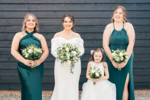 Four women stand in front of a dark wooden wall. The bride, in a white dress, holds a bouquet. Two bridesmaids in dark green dresses flank her, also holding bouquets. A young girl in a white dress smiles, holding a smaller bouquet.