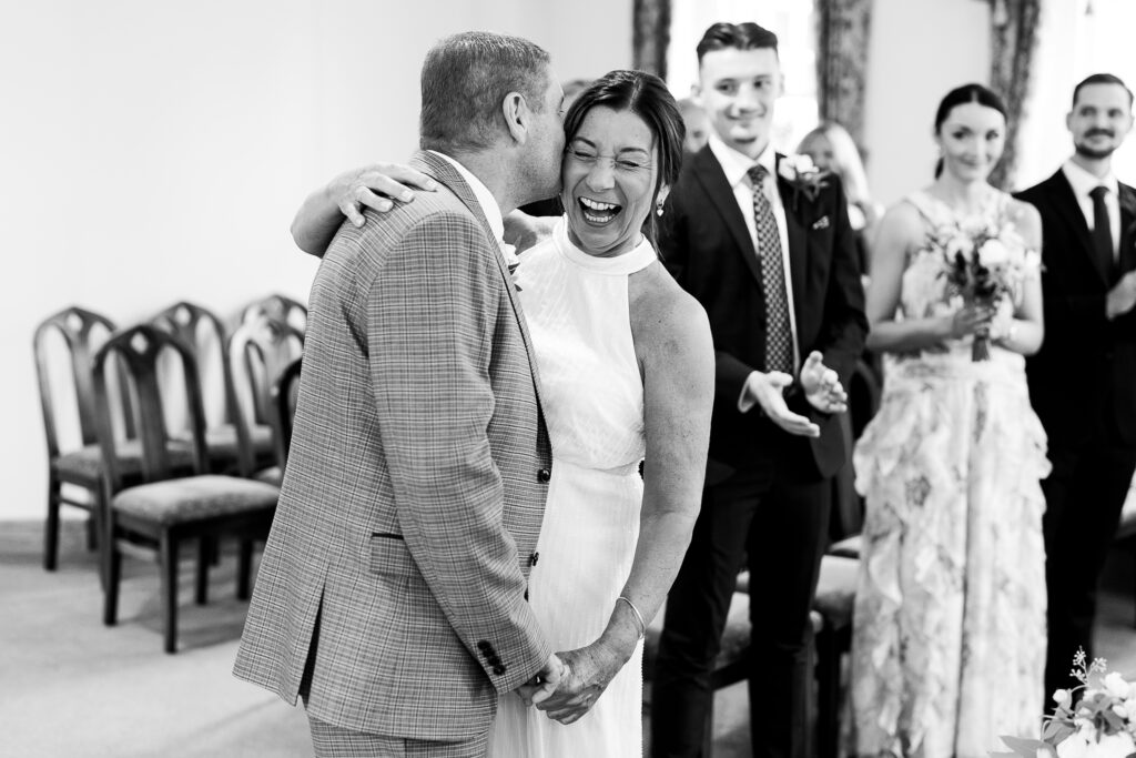 A bride and groom share a joyful moment, with the groom kissing the bride on the cheek. The bride laughs happily. In the background, guests applaud and smile. The scene is in a warmly lit room with chairs along the wall.