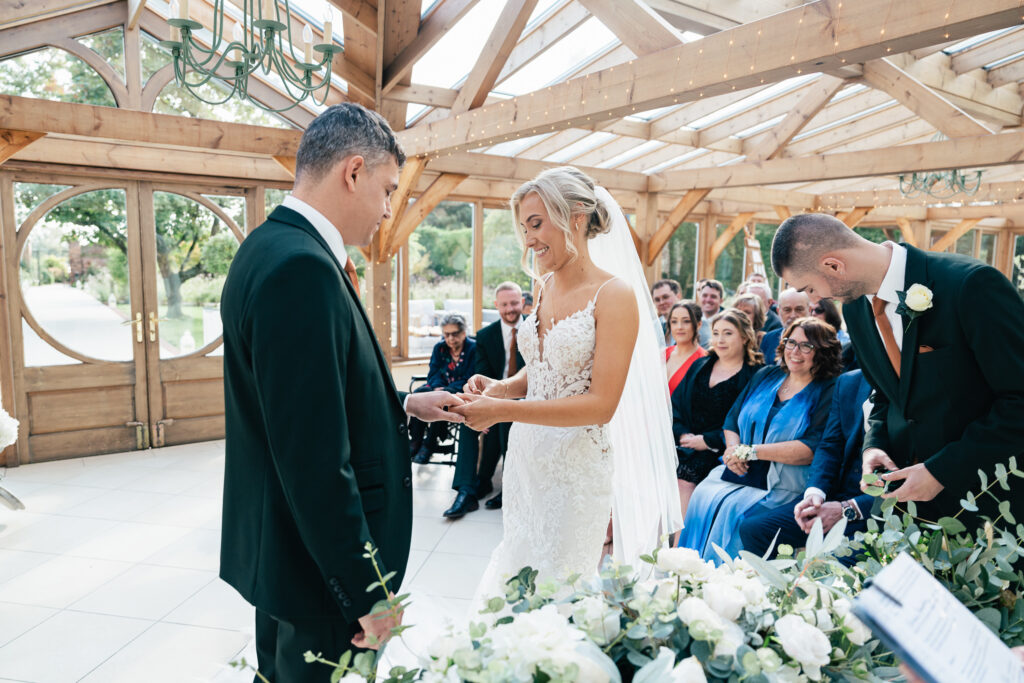 A bride and groom exchange rings in a bright, wood-framed venue with large windows. The bride wears a white lace gown, and the groom is in a black suit. Guests sit watching, surrounded by floral decorations. A groomsman stands beside them.