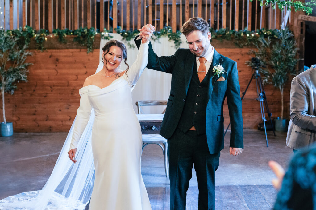 A bride in an off-the-shoulder white dress and a groom in a dark suit hold hands and smile. They are standing indoors, surrounded by greenery and wood paneling. A chair and blurred figures are visible in the background.