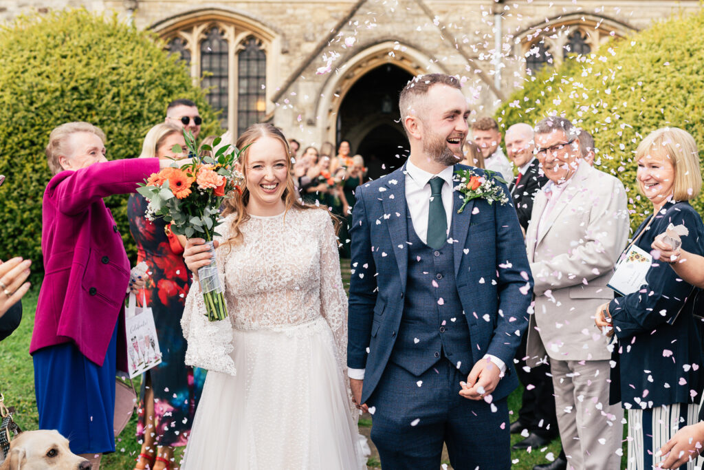 A bride and groom smile while walking down a confetti-strewn path outside a building with arched windows. The bride holds a bouquet, and people in formal attire stand on either side, celebrating their marriage.