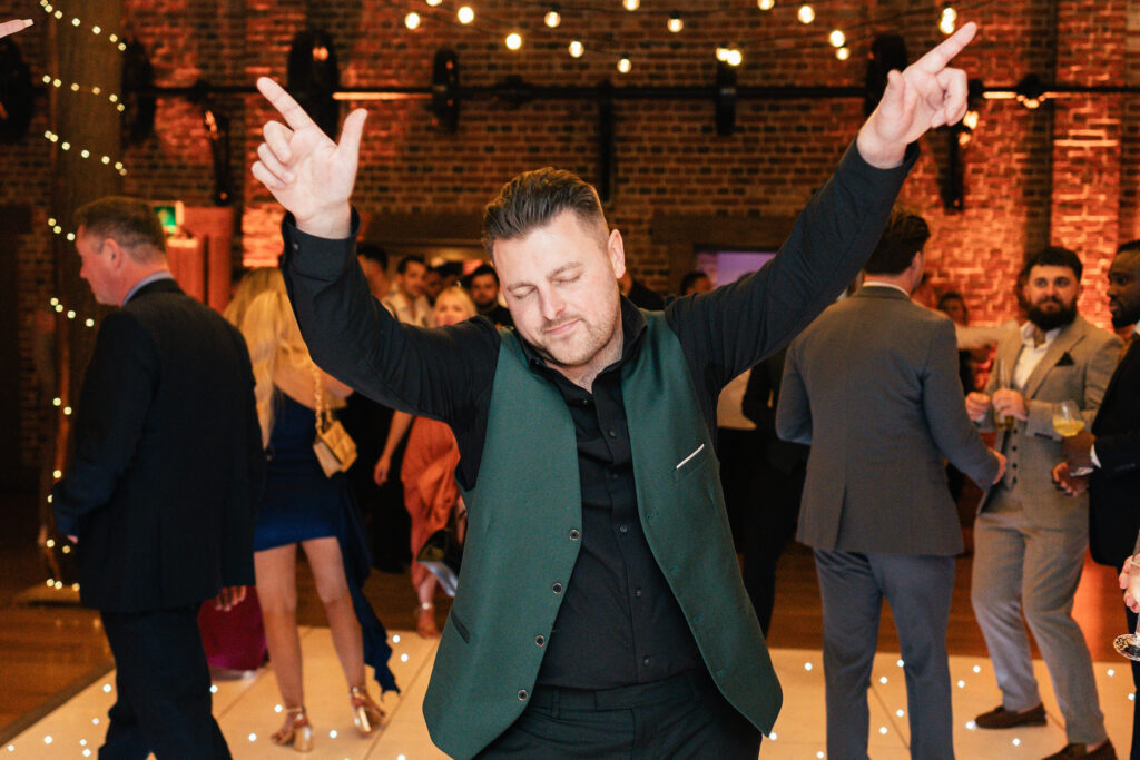 A person in a green vest and black shirt dances with both arms raised and eyes closed at a lively event. In the background, people socialize under string lights in a warmly lit, brick-walled venue.