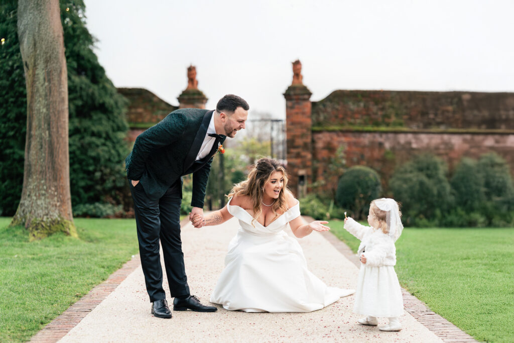 A bride and groom smile at a young girl in a white dress. The bride kneels, holding the girl's hand, while the groom stands beside them. They appear to be enjoying a moment together on a pathway in a garden setting.