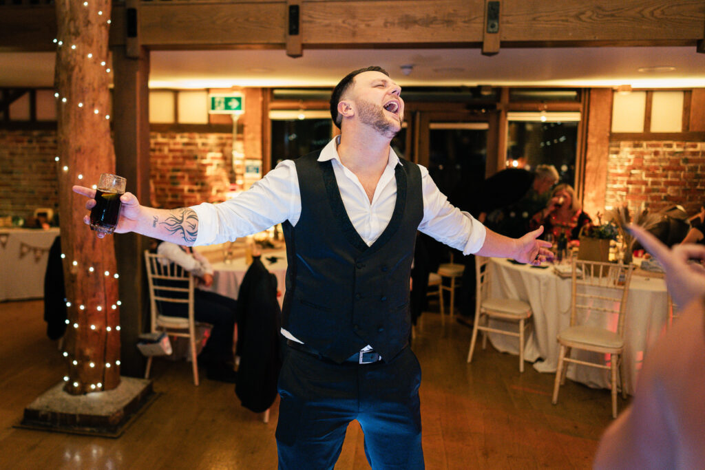 A man in a vest with rolled-up sleeves stands in a brightly lit room with brick walls. He holds a drink in one hand and joyfully sings or shouts, with his arms spread wide. Tables and chairs are in the background, and the setting appears festive.