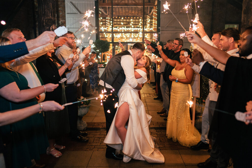 A bride and groom share a kiss as they are surrounded by guests holding sparklers. The bride wears a white dress with a high slit, and the groom is in a vest and pants. The scene is warmly lit with a festive backdrop.