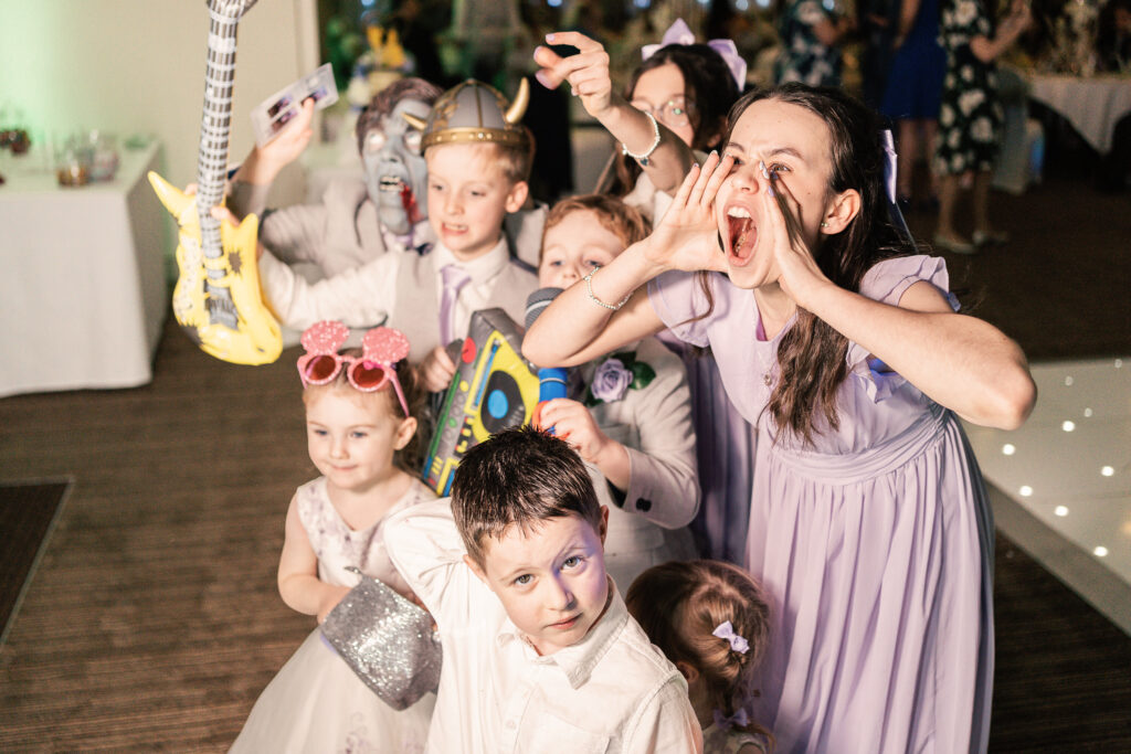 A group of children in playful outfits and props pose excitedly at an indoor event. One child shouts with cupped hands, while others hold inflatable instruments and wear fun accessories. The background shows a brightly lit room.