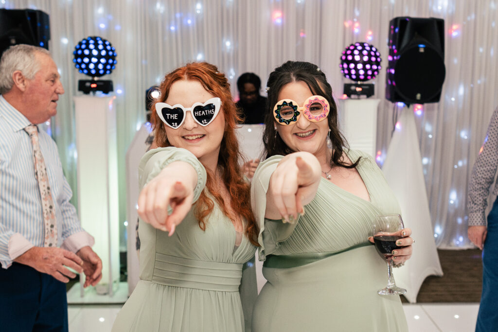 Two women in matching light green dresses wear novelty sunglasses and point towards the camera at a party. One has long red hair, and the other has dark hair and holds a wine glass. Colorful lights and guests are visible in the background.