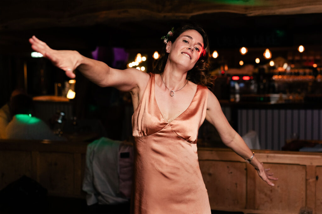 A woman in a peach satin dress dances joyfully, arms outstretched, in a dimly lit bar. She has a flower in her hair and appears to be enjoying the moment. The background features a bar with glowing lights and wooden decor.