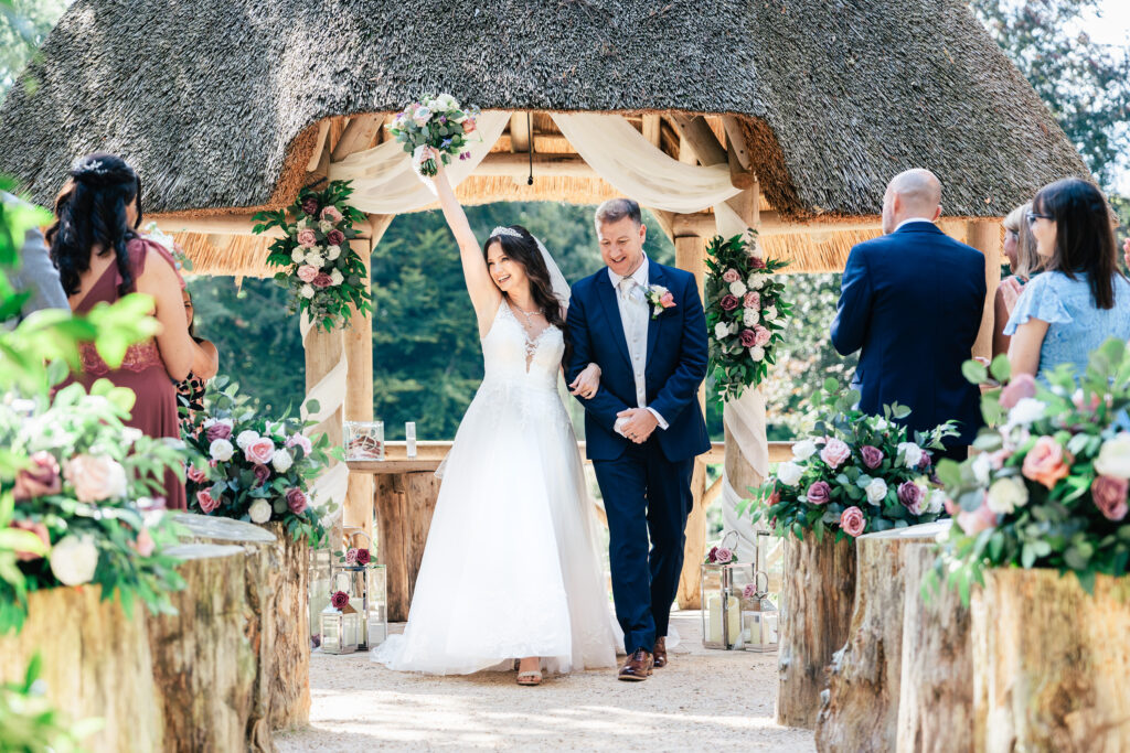 A bride and groom walk down an outdoor aisle under a thatched archway adorned with flowers. The bride raises her bouquet triumphantly. Guests stand applauding, surrounded by rustic decor with pink and white floral arrangements.