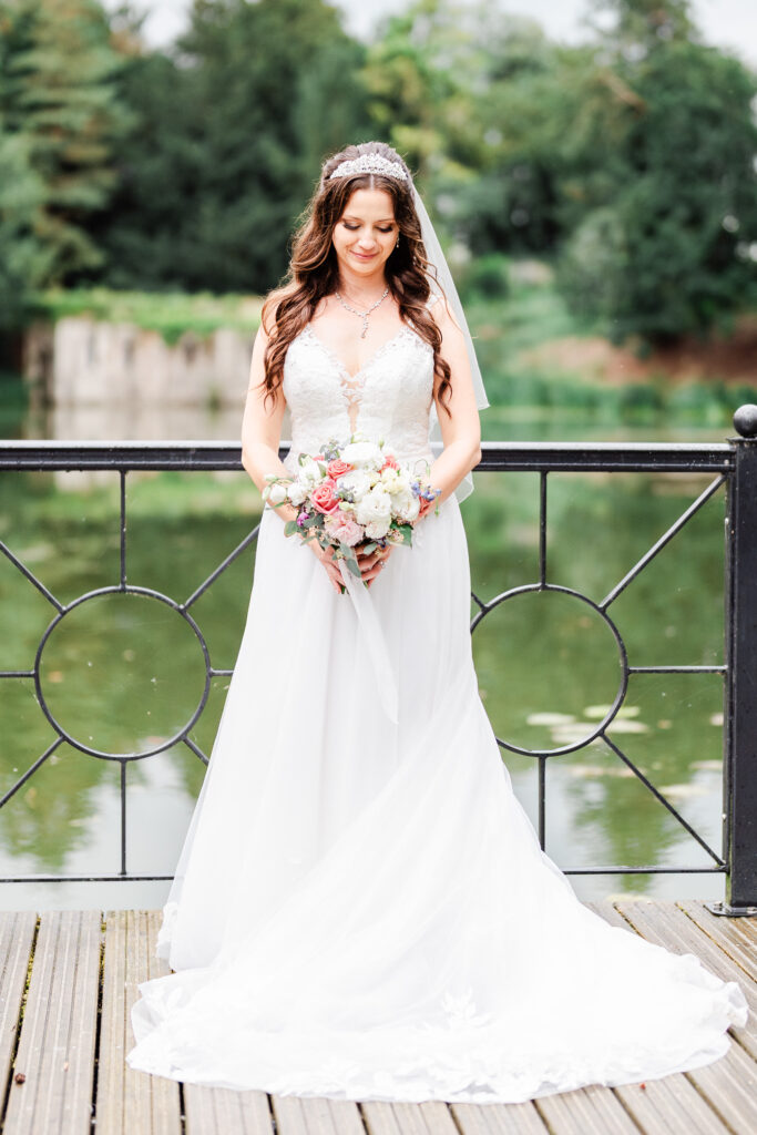 A bride stands on a wooden deck in front of a railing with circular designs, overlooking a lake. She wears a white wedding dress, holds a bouquet of flowers, and has a tiara on her head. Trees are visible in the background.