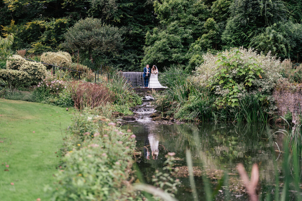 A couple stands on a small bridge over a stream, surrounded by lush greenery and a well-maintained garden. The reflection of the scenery is visible in the water below.