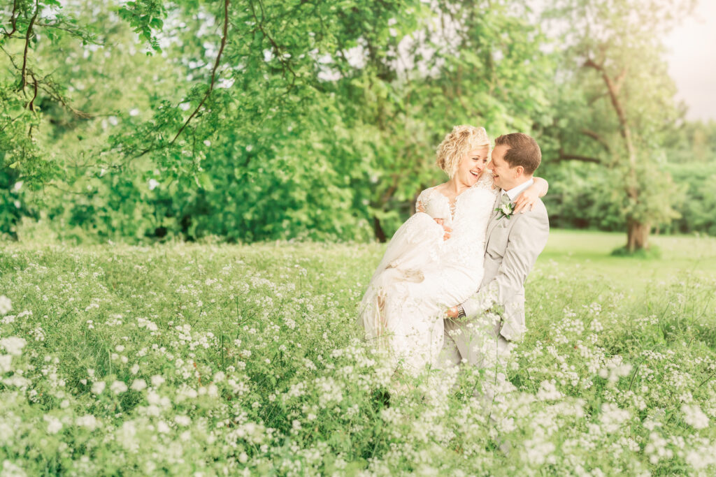 A groom in a gray suit joyfully carries his bride, who is wearing a white dress, through a field of white flowers. They are surrounded by lush green trees under a bright sky.