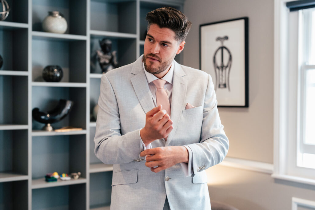 A man in a light gray suit with a pink tie and pocket square adjusts his cufflink in a modern room with gray shelving and framed art.