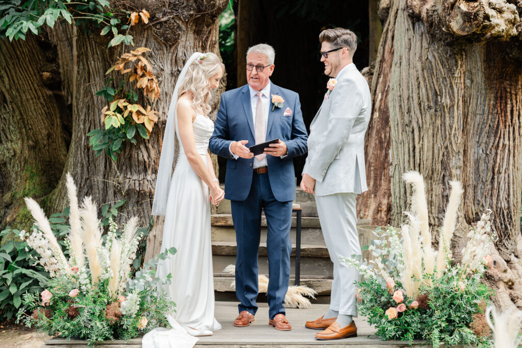 A couple stands on a wooden platform during their outdoor wedding ceremony. The bride is in a white dress with a veil, the groom in a light gray suit. The officiant, in a blue suit, holds a tablet. Rustic wooden backdrop with floral arrangements surrounds them.