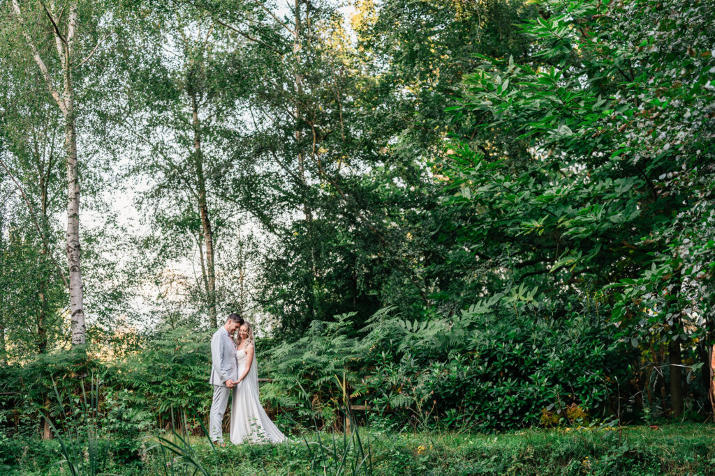 A newlywed couple stands closely together in a lush, green forest setting. Tall trees and dense foliage surround them, creating a serene, romantic atmosphere. The bride wears a white dress, and the groom is in a gray suit.