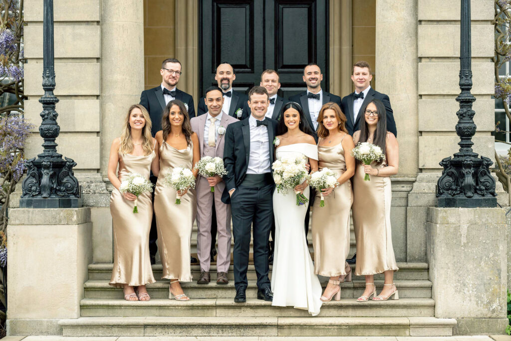 A wedding party stands on stone steps in front of an elegant building. The bride in a white dress is beside the groom in a tuxedo. Bridesmaids wear matching champagne dresses, and groomsmen are in tuxedos. Everyone is smiling and holding flowers.