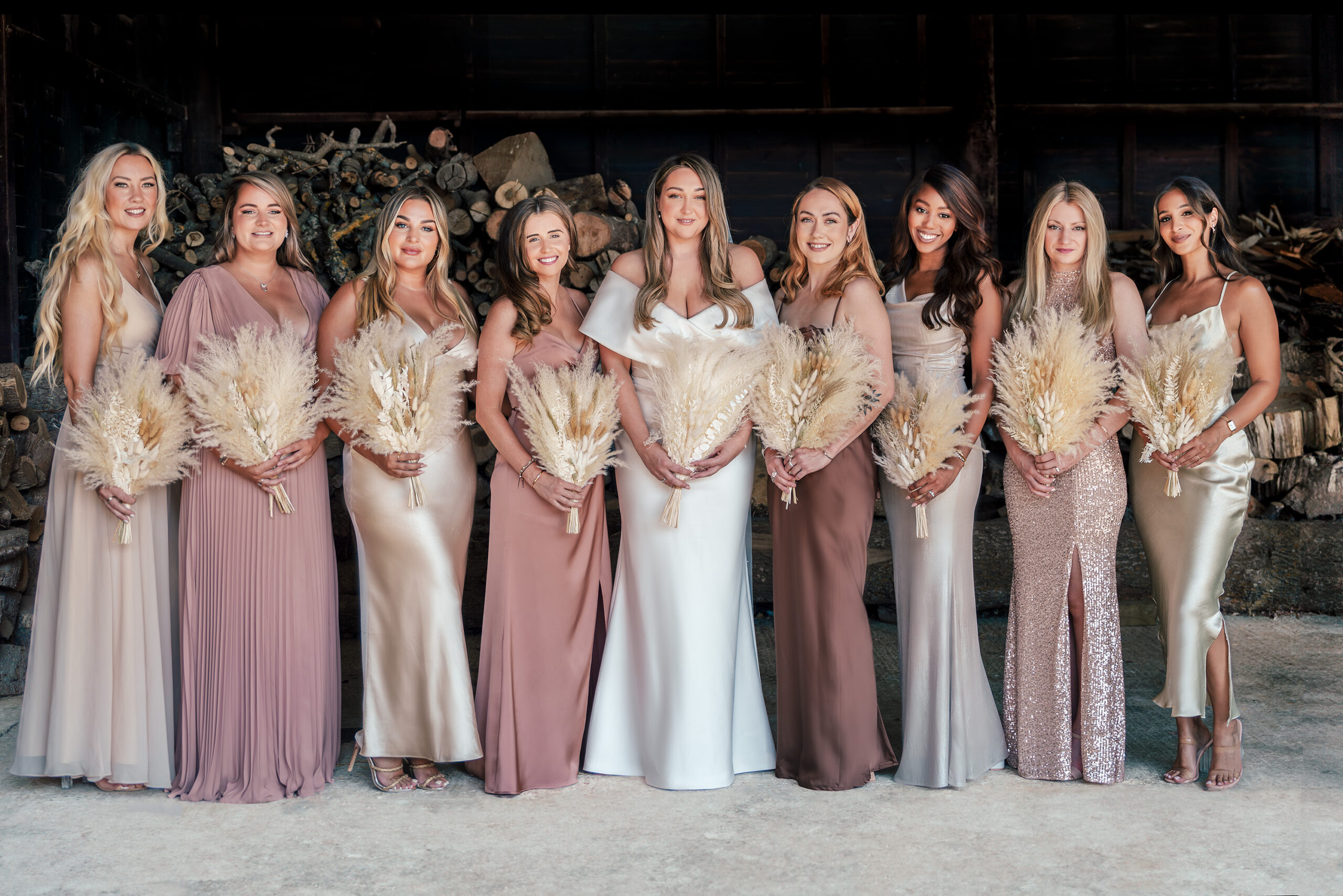 Nine women in elegant, neutral-toned dresses stand in a row, each holding a bouquet of pampas grass. They are posed against a rustic, wood-stacked background, smiling and facing the camera.