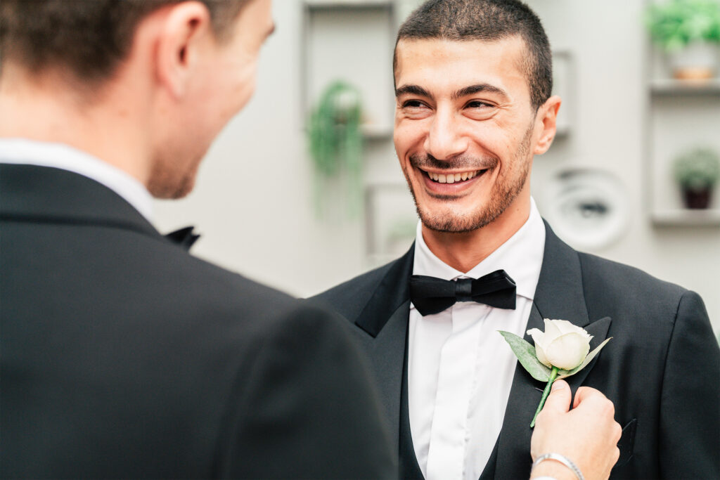 Two people in tuxedos smiling at each other. One person is pinning a white rose boutonniere on the other's jacket. The background is softly blurred with shelves and decorative items.