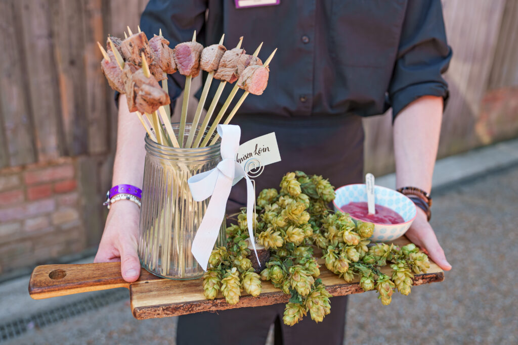 Person holding a platter with skewered meat in a jar, green hops, and a small bowl of dipping sauce. The platter is wooden, and the person is wearing a black outfit. A ribbon labeled "Dinner4u" is tied around the jar.