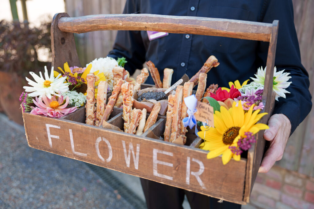 A person holds a rustic wooden flower box filled with assorted colorful flowers and crispy breadsticks. The box is labeled "FLOWER," adding a decorative and charming touch to the arrangement.