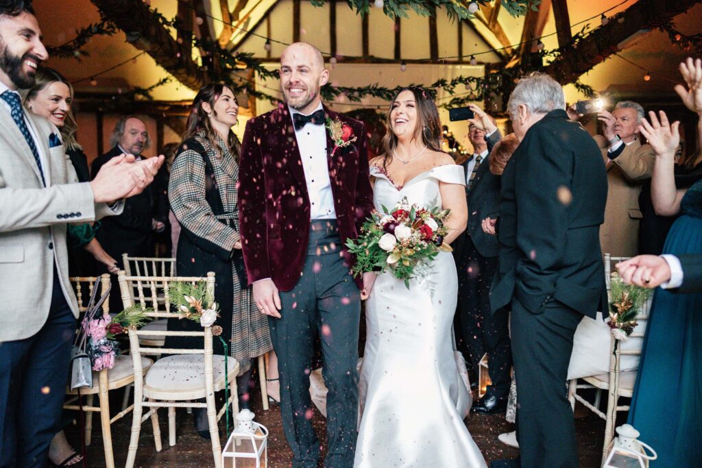 A bride and groom walk down the aisle amidst cheering guests. The groom wears a burgundy jacket, and the bride is in a white dress holding a bouquet. The venue is decorated with greenery, and guests are clapping and smiling.