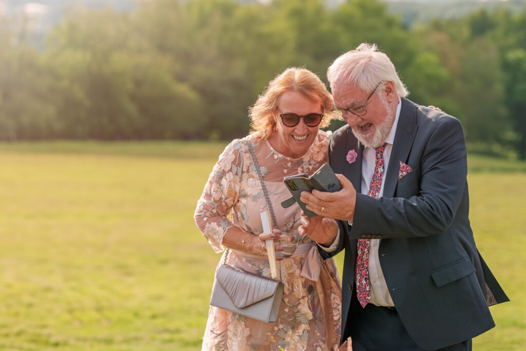 Elderly couple dressed in elegant attire, smiling and looking at a smartphone. They are standing outdoors in a grassy area with trees in the background, enjoying a sunny day.