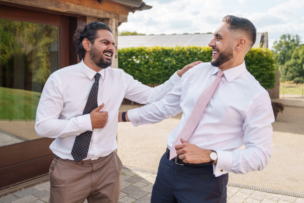 Two men in dress shirts and ties are outside, sharing a laugh. One has his arm around the other's shoulder. They appear to be in a cheerful, sunny setting with greenery in the background.