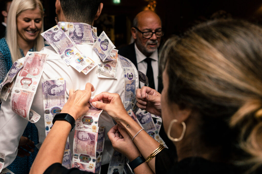 People are pinning banknotes to a man's white shirt at an indoor event. A smiling woman and a man in glasses are in the background. The scene conveys a festive or celebratory atmosphere.