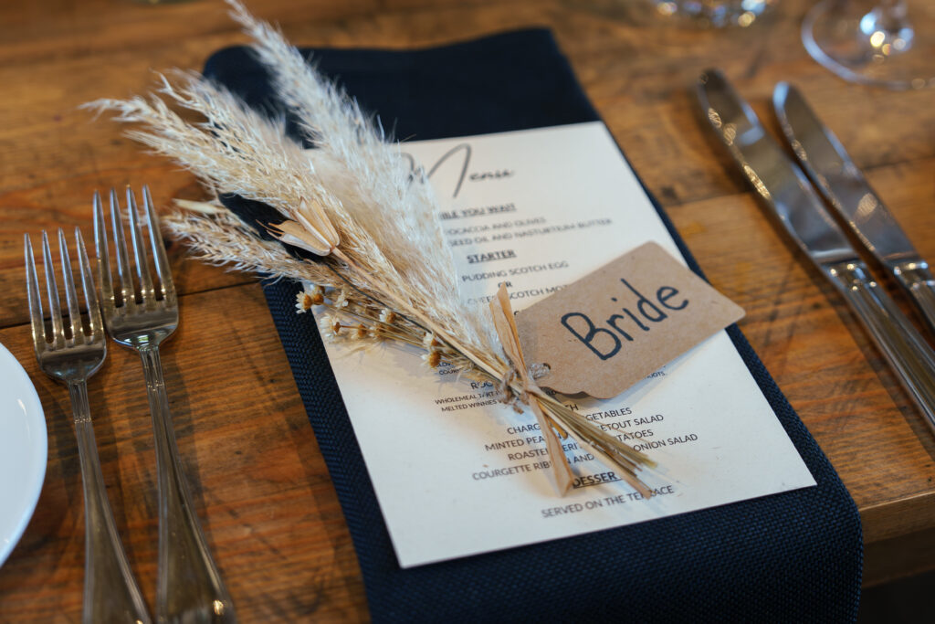 A wedding table setting features a menu on a dark blue napkin with a small bouquet of dried flowers tied with twine and a card labeled "Bride." Silverware is placed on either side on a rustic wooden table.