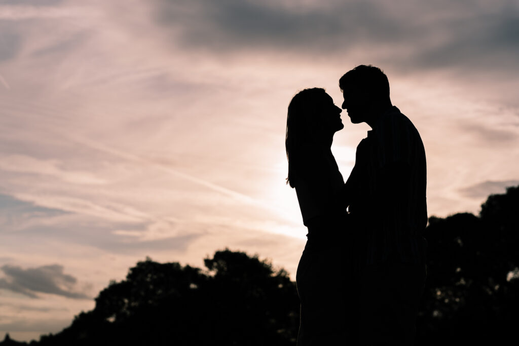 Silhouette of a couple about to kiss against a dramatic sunset sky, with clouds and tree outlines in the background.