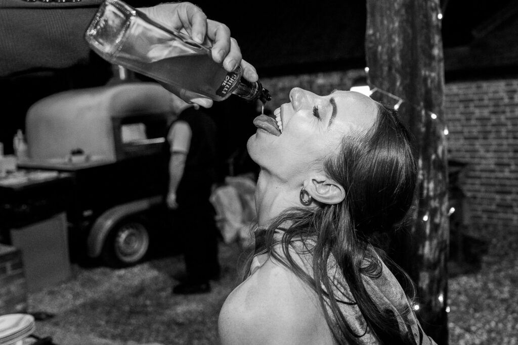 A woman with long hair tilts her head back, laughing, as someone pours liquid from a bottle into her mouth at a nighttime outdoor gathering. There's a food truck in the background and string lights around a wooden post.