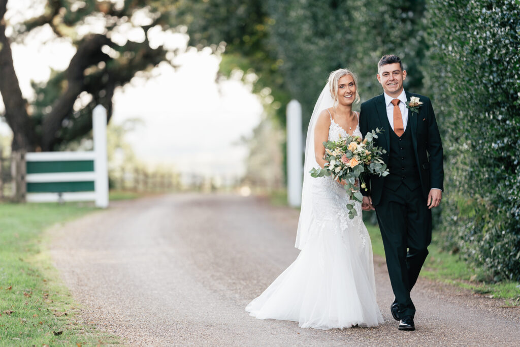 A bride in a white dress and veil holding a bouquet of flowers walks arm-in-arm with a groom in a black suit and orange tie. They are smiling, walking down a tree-lined path with a sunny background.