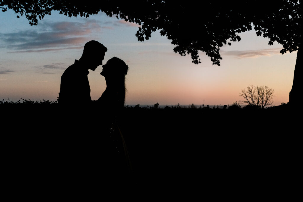 Silhouette of a couple standing close under a tree, set against a colorful sunset sky with patches of clouds and distant trees on the horizon.