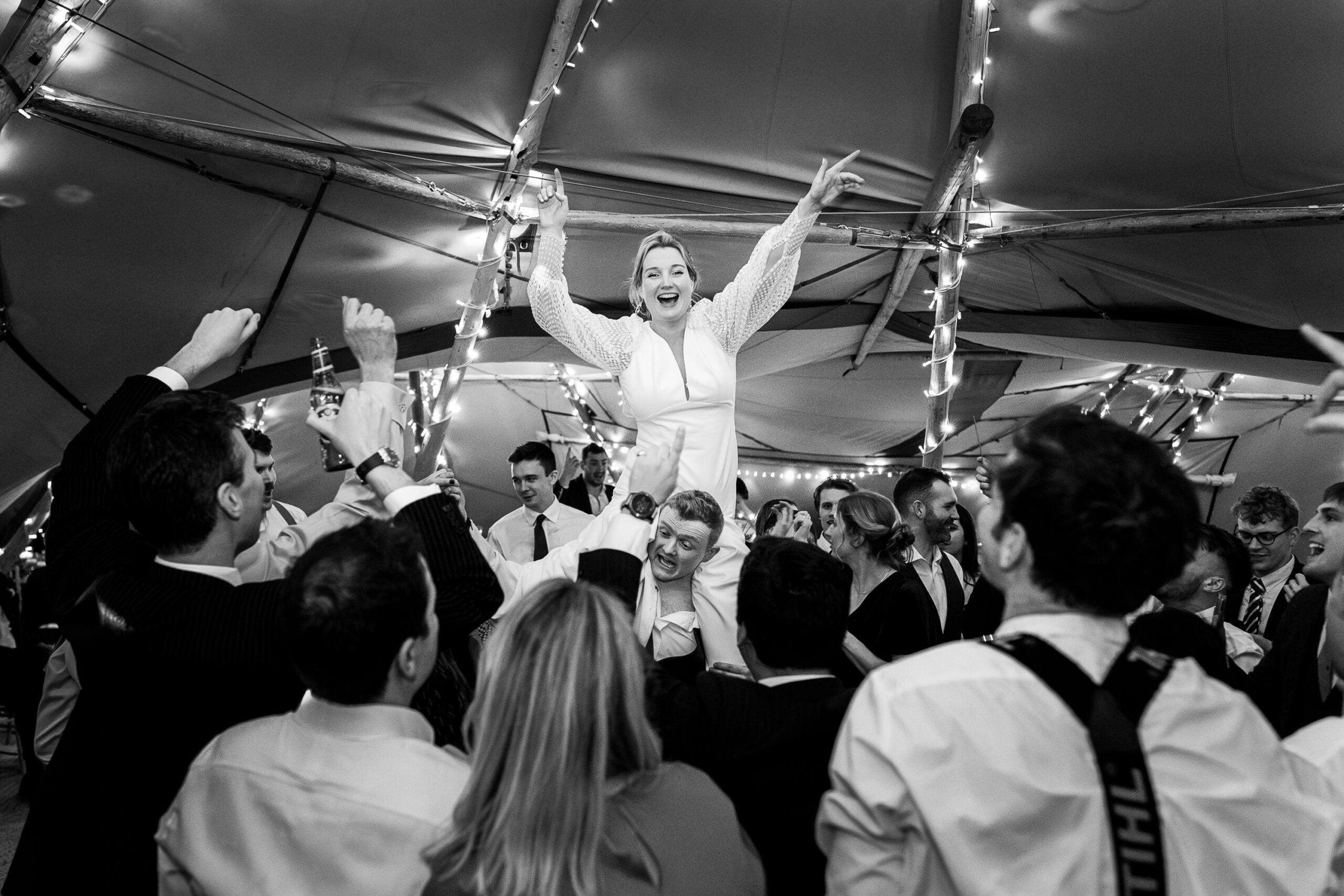 A black and white photo of a joyful wedding celebration. The bride, smiling broadly, is lifted into the air by a crowd of enthusiastic guests under a tent decorated with string lights. People around are cheering and raising their hands in excitement.