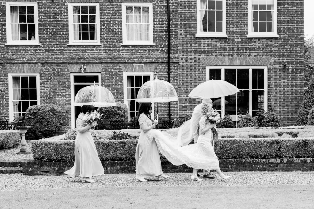 Black and white image of a bride in a wedding dress walking in front of a brick building, followed by three bridesmaids. Each holds a transparent umbrella, and one bridesmaid holds the bride's train. They walk along a garden path.