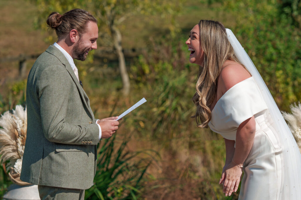 A bride and groom stand outdoors, exchanging vows. The groom, in a light gray suit, smiles while reading from a paper. The bride, wearing a white off-shoulder dress and veil, leans forward, laughing joyfully. The background is a sunny, green landscape.