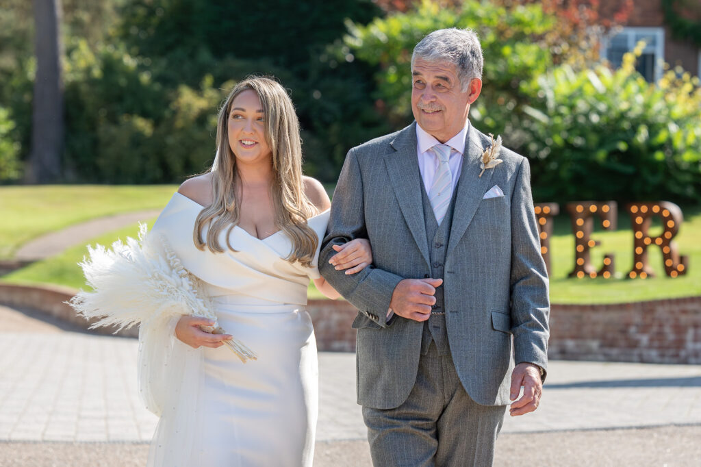 A bride in a white dress walks arm-in-arm with a man in a gray suit. She holds a bouquet of white flowers. They appear to be outdoors, with greenery and a large sign in the background.