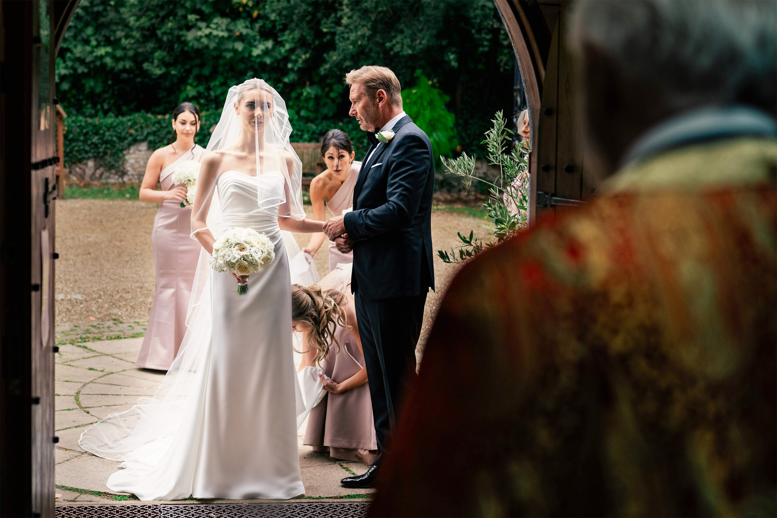 A bride in a white dress and veil holds flowers, standing next to a man in a suit at the entrance of a building. Bridesmaids in light dresses adjust her gown as an officiant, in ornate robes, stands nearby. Lush greenery is visible outside.