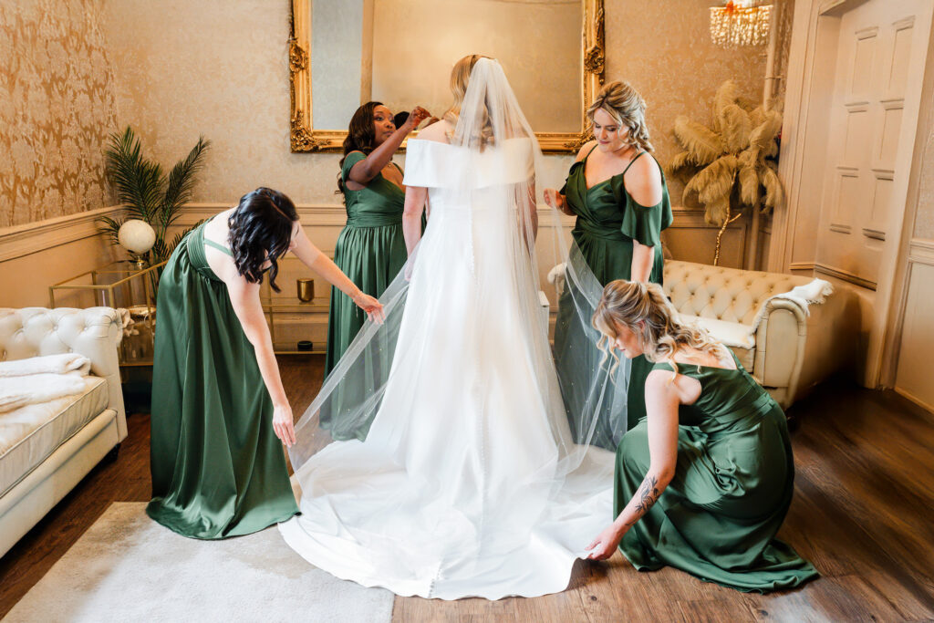 A bride in a white gown and veil stands as four bridesmaids in green dresses help arrange her dress in an elegant, softly lit room with cream-colored furniture and ornate decor.