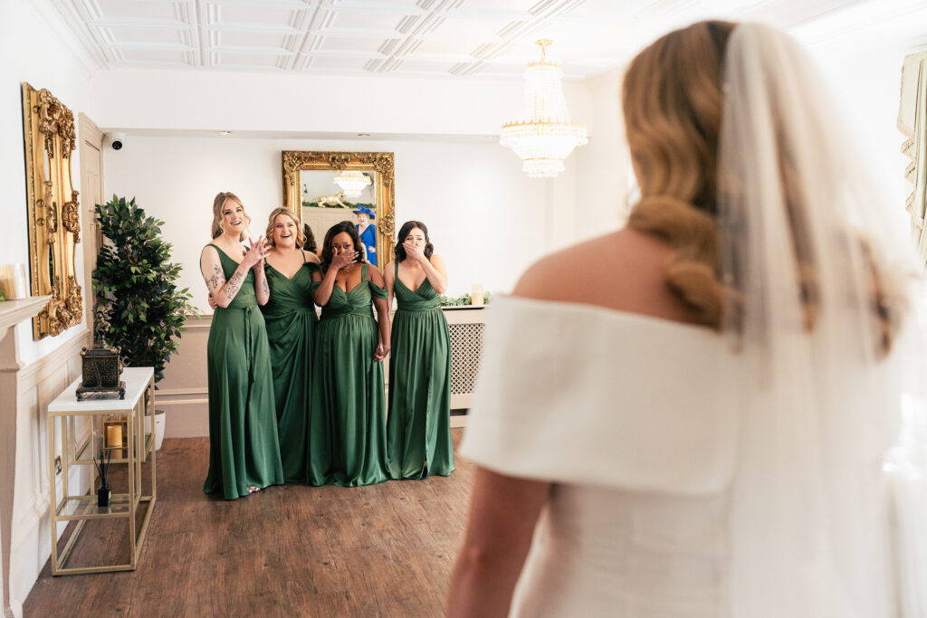 Four bridesmaids in green dresses react with joy and surprise as they see the bride, who is facing them in a white dress and veil, in a bright, elegant room with a chandelier and decorative mirror.